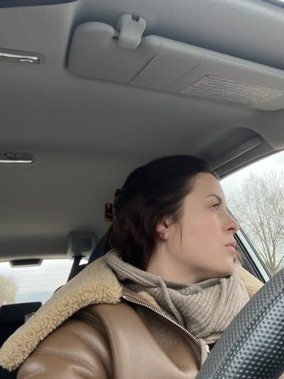 A female individual is depicted in the passenger seat of a vehicle, located in Schmittweiler, Germany. The subject is shown in profile, looking towards her right through the vehicle's window. She has dark hair, partially secured by a tortoiseshell-patterned claw clip, and a light complexion. Her attire consists of a brown faux leather jacket with a cream-colored faux shearling collar, layered over a beige ribbed knit snood covering a hooded sweatshirt.

The vehicle's interior features a light grey headliner. A light grey passenger-side sun visor is in its upright position, displaying a warning label on its underside. The top portion of a black, textured steering wheel is visible on the right side of the frame. Outside the vehicle, a view through the window reveals bare deciduous trees and an overcast sky, suggesting cool weather. Distant, indistinct structures are faintly visible on the horizon.