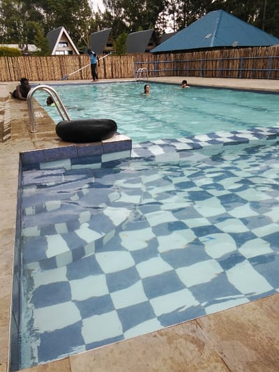 A pool scene reveals several individuals: three people swimming in the main pool, and a man lounging at the pool's edge near a black inner tube. Another man stands nearby, seemingly cleaning the pool. The pool features a shallow section with blue and white checkered tiles, adding a decorative element. The background shows buildings with peaked roofs, surrounded by a bamboo fence and trees, suggesting a resort or recreational area.