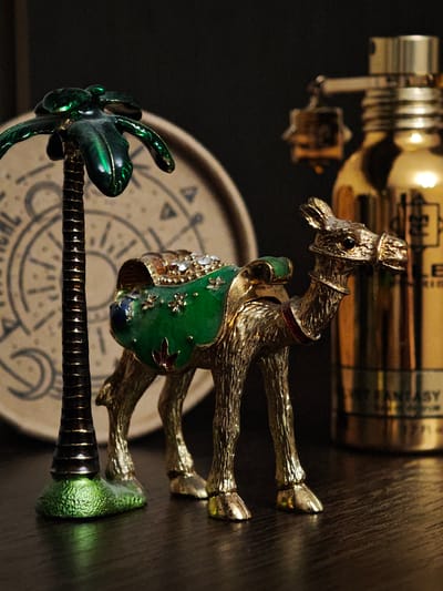 An indoor, eye-level, close-up photograph shows a gold-colored decorative camel figurine with a green saddle adorned with rhinestones and enamel. The camel is standing on a dark wooden surface. To the left of the camel is a miniature green palm tree figurine with a textured trunk. In the background, slightly out of focus, is a circular object with a celestial or astrological pattern and a tall, golden bottle, possibly perfume, with some text visible. The lighting is dim, casting shadows and highlighting the metallic sheen of the figurines. The scene appears to be a tabletop arrangement.