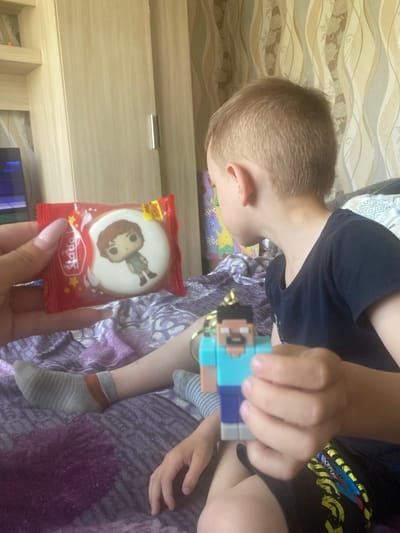 The image shows a person's hands holding a small, white, round cookie with a picture of a cartoon character on it, which is packaged in red foil. The cookie package has text that appears to say "Sadovaya" and some yellow stars. In the background, a child with blonde hair, wearing a dark t-shirt, is holding a small, multi-colored toy, possibly a keychain. The child is sitting on a bed covered with a purple blanket. There are also some colorful patterned fabrics and a wooden wall visible in the background. The lighting suggests it could be daytime indoors.