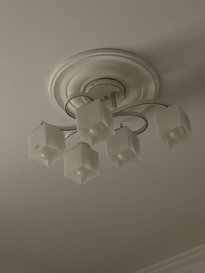 This media file shows a close-up, slightly tilted-down view of a ceiling light fixture. The fixture has a decorative, circular base mounted on a white ceiling. Attached to the base are several arms that hold individual lamps. Each lamp is enclosed in a frosted, hexagonal-shaped shade with a ribbed texture. Inside each shade, a light bulb is visible. The overall scene suggests an indoor setting, likely a room in a residential or commercial building. The lighting appears to be artificial, as the light bulbs in the fixture are illuminated. There are no people visible in the media file. The focus is entirely on the lighting fixture itself and the surrounding ceiling. The angle of the shot gives a sense of looking up at the fixture. The colors are muted, primarily white and off-white from the fixture and ceiling, with some darker tones from shadows and the metallic parts of the fixture. The ribbed texture of the lampshades is a notable detail, creating a pattern of light and shadow.