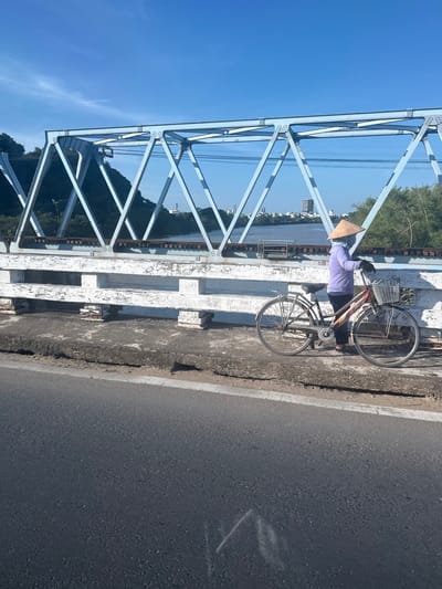 A person wearing a conical hat and a light purple shirt stands next to a bicycle on a bridge. The bicycle has a basket on the front. The person appears to be leaning on the handlebars and looking to the right. The bridge has a metal structure on the left side and a concrete railing on the right. In the background, a river or body of water is visible, with trees and buildings on the far bank. The sky is clear and blue, suggesting a sunny day. The overall scene depicts a person pausing on a bridge, possibly taking a break from cycling. The time of day appears to be daytime, given the bright sunlight and clear sky. The location, Tuy Hoa, Vietnam, suggests a warm climate.
