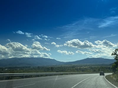 The image captures a winding asphalt road with white lane markings, leading through a rural landscape under a bright blue sky dotted with white cumulus clouds. A silver guardrail runs along the left side of the road. In the distance, rolling hills and mountains form the horizon. A dark-colored SUV is visible driving away on the road, taking the curve to the right. The foreground features the road surface, while the middle ground shows a verdant expanse of trees and vegetation. The background is dominated by the mountain range. The overall impression is one of travel and a scenic, open-air environment. The image is likely taken in or around Konyavo, Bulgaria, based on the geographical context provided.