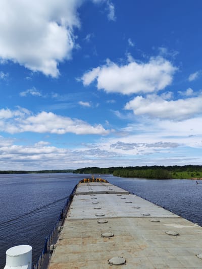 The image depicts a view from the deck of a flat-top barge on a wide body of water, possibly a river or lake, in Pruzhinino, Russia. The barge has a metal deck with evenly spaced circular access hatches. Blue railings line the edges of the barge. The water is dark blue and has visible ripples, indicating movement. On the right bank, there is a line of green trees and some open grassy areas. The sky is bright blue with numerous fluffy white clouds.