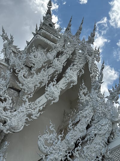 The image shows a close-up, low-angle view of the ornate white facade of the Wat Rong Khun, also known as the White Temple, in Ban Rong Khun, Thailand. The temple's architecture is characterized by intricate, swirling white stucco decorations, likely incorporating mirrored elements that reflect light, against a background of a partly cloudy blue sky. The details visible are primarily architectural, focusing on the elaborate patterns and spires that adorn the temple's roofline and eaves. No people are visible in the frame. The environment is an exterior setting, with the sky serving as the backdrop.