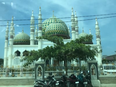 The image shows a white mosque with green and gold domes and tall minarets against a blue sky, partially obscured by a tree in the foreground. Several motorcycles are parked in front of a fence that surrounds the mosque. The location appears to be in an urban setting, as suggested by the presence of vehicles and buildings in the background. The location is Rawang Itiek, Indonesia.