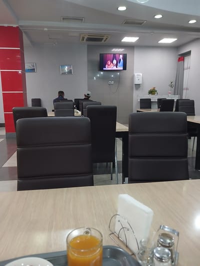 A medium shot captures the interior of a canteen or cafe, potentially located in Kotino, Russia. The scene is dominated by tables and chairs, with a television screen mounted on the wall showing cartoon characters. The chairs are upholstered in dark leather and arranged around light-colored wooden tables. In the foreground, a tray holds a glass of orange juice, a napkin dispenser, and salt and pepper shakers. Two individuals are visible in the background, seated at tables and facing away from the camera, their identities obscured. The walls are painted a neutral grey, with red accents on a pillar to the left. The lighting appears to be artificial, with recessed ceiling lights and spotlights illuminating the space. The overall impression is one of a functional, everyday dining establishment.