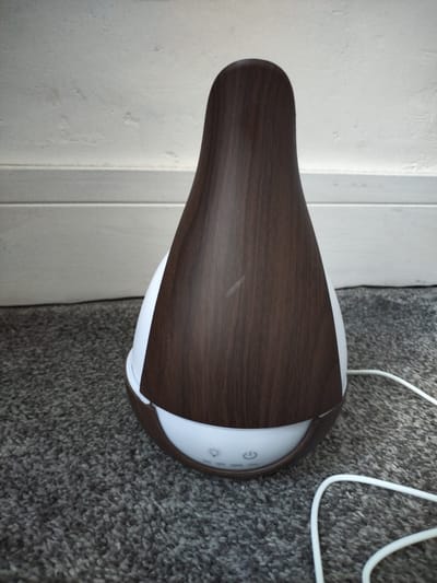 A small, white and dark wood-grain essential oil diffuser sits on a grey carpet. The diffuser has a teardrop shape with a dark wood-grain exterior and a white interior. The base of the diffuser has three illuminated buttons. A white power cord extends from the right side of the diffuser and is visible on the carpet. The background consists of a light-colored wall with white baseboards. The scene is indoors, with no specific location in Darlington, United Kingdom, identifiable from the image.
