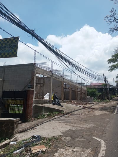 The image depicts a street scene in what appears to be a residential or commercial area. An unfinished building constructed of concrete blocks is visible on the left side, indicating ongoing construction. Wood framing is in place, along with vertical reinforcing steel. The front of the building has a sign that says "CLEAN GLOW CAR WASH." A black metal gate is partially visible in front of the building. A pile of refuse is in the foreground. Utility poles and wires span the street, running diagonally across the frame. The road surface is cracked and shows signs of wear. Vegetation grows along the sides of the road and in between cracks in the pavement. Other buildings are seen in the background, including one with a red tile roof. The location is Kioumaknao 2, Laos. The weather appears sunny with some clouds. There are no people visible in the image.