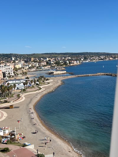 This image depicts an elevated view of a coastal area within Civitavecchia, Italy, under a clear blue sky. The upper approximately 20% of the frame shows a cloudless sky, with a single faint white cloud appearing horizontally near the left-center horizon.

The mid-ground features the city of Civitavecchia, characterized by a dense arrangement of light-colored buildings with scattered terracotta roofs, extending along the coastline and ascending into green, undulating hills in the background. A prominent white structure featuring multiple arches is visible extending into the water from the urban area on the middle-left. Adjacent to this, a harbor area is protected by a long, rocky breakwater that curves eastward into the sea. Small figures are visible walking on this breakwater.

The foreground is dominated by a wide, crescent-shaped pebble beach that transitions from a paved promenade and associated structures on the left to the Tyrrhenian Sea on the right. The water near the shore is a light turquoise, deepening to a dark blue further out. Gentle waves are present along the waterline. Two small white sailboats are discernible in the darker blue water in the mid-right.

Approximately 20-30 individuals are present on the beach. Several persons are observed reclining on towels or beach chairs on the dry sand. Multiple small groups, typically comprising 2-5 individuals, are standing or walking near the water's edge and on the main beach area. On the lower-right section of the beach, a group of approximately five individuals is standing, with a small white object, possibly a ball, near their feet, suggesting recreational activity. A single individual is partially visible on a paved area in the far lower-right, with an arm extended.

Along the promenade behind the beach, there are paved walkways, green landscaped areas, and several palm trees. A multi-story white building with blue accents and multiple palm trees is situated directly behind the left portion of the beach. The right edge of the frame is partially obscured by a vertical white architectural element.