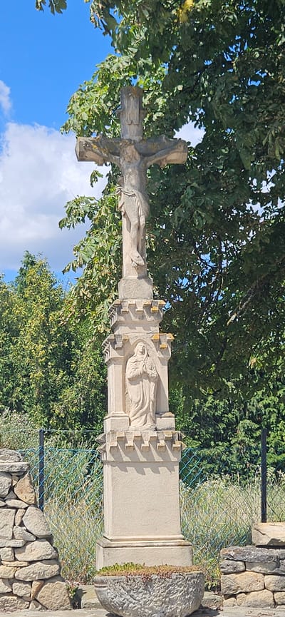 An outdoor, eye-level, full shot captures a stone crucifix in Cserszegtomaj, Hungary, set against a backdrop of lush green trees and a bright blue sky with scattered clouds. The crucifix, made of light-colored stone, stands on a multi-tiered pedestal. The figure of Jesus is depicted on the cross, and below it, on the pedestal, is a carved relief of a praying woman in a habit. The base of the pedestal is adorned with a decorative crenellated pattern. In the foreground, a rustic stone wall and a wire fence partially enclose the scene. A weathered stone planter filled with small green plants sits at the base of the pedestal. The overall impression is one of historical and religious significance in a natural setting.