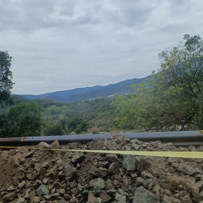 The image captures a scenic view of rolling hills covered in dense green trees under a cloudy sky. In the foreground, a pile of rocks and dirt is visible, with a rusty pipe running horizontally above it, marked by a yellow caution tape. The scene is located in Madzharovo, Bulgaria, a region known for its natural beauty and mountainous terrain.