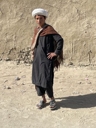 The image depicts a young male standing outdoors in Spin Boldak, Afghanistan. He is positioned centrally, facing slightly left of the viewer with a neutral expression. He wears a white turban, a brown and beige striped shawl draped over his shoulders, a black long-sleeved tunic, dark pants, and grey open-toed sandals. His left hand rests on his hip. Behind him is a tall, textured wall composed of dried mud or earth, displaying numerous cracks and eroded surfaces. Faint, dark script is partially visible on the upper left portion of the wall. The ground beneath is an unpaved, light brown dirt surface, marked with footprints and scattered debris including small pieces of light-colored refuse and a pink item. A defined shadow of the individual extends to the right on the ground, indicating direct overhead sunlight.