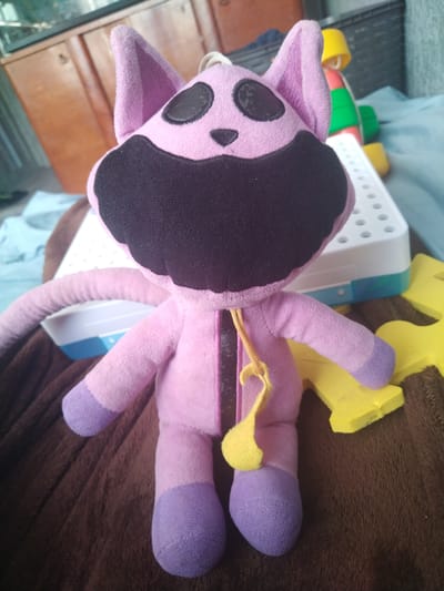 A pink plush toy with purple paws and ears is lying on a brown blanket. The toy has a large black mouth and black oval eyes. A yellow string hangs from the toy's neck. Behind the toy is a white and blue object with many small holes, and to the right, a yellow foam cutout in the shape of an "H". In the background, a wooden cabinet and a portion of a blue object are visible. The scene appears to be indoors.