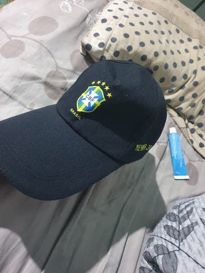 The image shows a close-up of a black baseball cap with the Brazilian national football team logo and the word "BRASIL" embroidered on the front. The cap also has the name "NEYMAR JR" embroidered on the side. A blue tube of what appears to be toothpaste or ointment is lying next to the cap on a surface covered with a patterned fabric. The background appears to be a bed or sofa with a dotted pillow. There are no people visible in the image. The location is not discernible from the image provided; therefore, its presence in Ghafurov, Tajikistan cannot be confirmed.