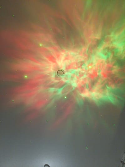 The image displays a projected nebula-like pattern of red and green lights on a ceiling. Scattered green dots, resembling stars, are also visible across the ceiling. In the center of the projected nebula, there is a circular fixture. The scene appears to be an indoor setting, likely a room, where a projector is casting these lights onto the ceiling. There are no people, animals, or any outdoor elements depicted. The specific location in Vasilievo, Russia, is not discernible from the visual information provided in the image.