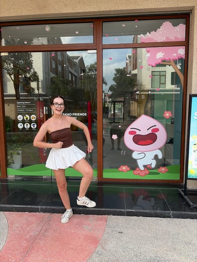 A young woman with glasses and brown tube top and a white skirt stands in front of a store window displaying a cartoon character. The store window has a depiction of a pink tree with falling blossoms. The woman is smiling and striking a pose with her hands on her hips. The store appears to be a Kakao Friends store, indicated by the text on the window. There is also a sign with opening hours and icons, including "NO FOOD," "NO SMOKE," and "NO PET." The exterior of the building is beige, and the sidewalk is made of pinkish concrete. The scene is outdoors, with reflections of buildings and trees visible in the store window. The image is set in Nghĩa Trụ Commune, Vietnam.