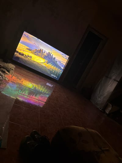 The image shows an interior scene, likely a residential room, with a large flat-screen television displaying a landscape of rolling hills and cypress trees, characteristic of Tuscany, Italy. The television screen also displays a timestamp "00:02 ч.", indicating it is likely a video or live broadcast. The room is dimly lit, with the primary light source emanating from the television.
The floor is tiled with a reddish-brown marble pattern and reflects the colorful light from the screen, creating a rainbow effect. In the foreground, to the lower left, a pair of dark shoes is visible, suggesting someone is seated or lying down. To the right foreground, a person's legs and lower torso are visible, partially obscured by shadow and a light-colored, possibly fabric-covered object with dark markings.
In the background, to the right of the television, there is an open doorway leading to a dark, unlit space. To the left of the television, there is a pile of what appears to be discarded plastic bags and papers. Further back on the right, there is a large object covered in white plastic sheeting, possibly furniture or storage.
The overall impression is a relaxed, perhaps evening, scene where someone is watching television in a somewhat cluttered or in-progress living space. The location in Shishmantsi, Bulgaria is not visually evident from the image itself.