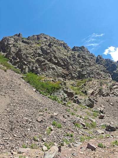 A rocky mountain slope under a clear blue sky with a few wispy clouds. The slope is covered in loose stones and gravel, with sparse green vegetation growing in patches. The mountain itself is rugged and steep, with visible rock formations and crevices. The scene is devoid of people and any man-made structures.