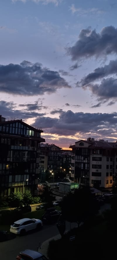 The image depicts a twilight scene in the city of Romashkovo, Russia, looking upwards towards the sky. The sky is filled with dramatic, dark, and grey clouds, with streaks of pastel pink and orange visible near the horizon, indicating the setting or rising sun. Below the sky, multi-story apartment buildings are visible, characterized by dark facades with numerous windows. The buildings have distinctive sloped roofs with balconies and railings. Streetlights are illuminated, casting a warm glow on the dark asphalt roads and sidewalks. Several cars are parked along the street and in parking areas, including a white SUV in the foreground. Lush green trees and foliage are interspersed throughout the scene, adding a touch of nature to the urban environment. The overall atmosphere is serene and peaceful, captured during the transitional light of dusk.