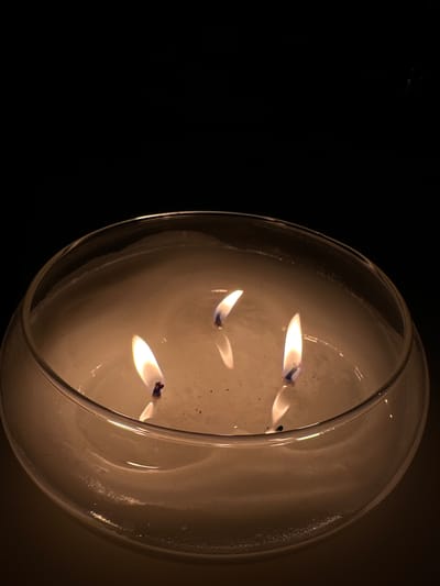 The image shows a close-up of a three-wick candle burning in a clear glass bowl. The candle is white and appears to be made of wax. The flames are a warm yellow and orange color, and they are casting a soft glow on the wax. The background is dark and out of focus. There is no indication of the location in the city of Rassokha, Russia.