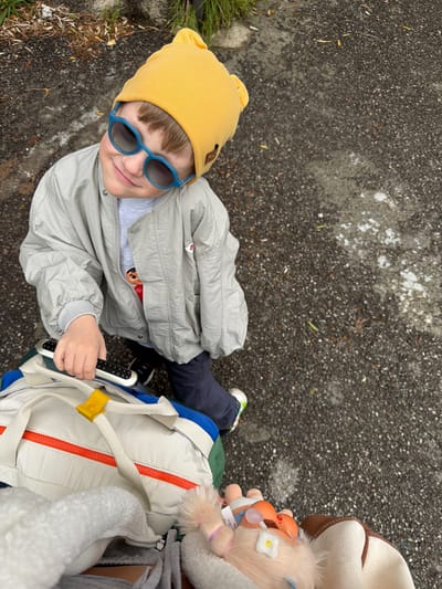 An overhead shot captures a young child looking up at the camera with a bright smile, outdoors in Nerin, Montenegro. The child, likely a toddler or preschooler, wears a vibrant yellow beanie with bear ears and stylish blue hexagonal sunglasses. They are dressed in a light gray jacket, dark blue pants, and athletic shoes (partially visible).
The child holds a silver and black remote control-like device in their right hand. Next to them, a light-colored backpack with a prominent orange zipper, blue, and green sections, and a yellow strap accent is visible. Two small plush toys are attached to the backpack or a nearby person: one resembles a baby doll, and the other is a fluffy animal adorned with a small white flower. A small, unreadable white tag with blue text is visible on the baby doll plush. Part of an adult's attire, including a brown and white hat and a white fluffy garment, is seen on the far right, suggesting an adult is accompanying the child.
The setting is an outdoor paved area, likely asphalt, speckled with white and a few scattered dry leaves. Patches of green grass and dirt are visible along the left edge. The diffused lighting indicates an overcast day. The child's happy expression and engaging gaze are notable.