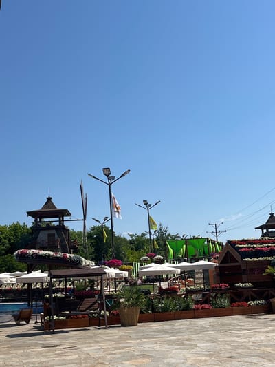 The image captures an outdoor scene in Gorna Malina, Bulgaria, likely a park or recreational area. A bright blue sky dominates the upper portion of the frame. In the middle ground, a rustic, multi-story wooden structure with a peaked roof and balcony stands prominently, flanked by trees. Several streetlights with multiple LED panels are visible, some equipped with flags. To the right, a large wooden structure resembling a house or a tiered display is covered in a vibrant array of red and white flowers.
In the foreground, a paved area with large, irregular flagstones leads into the scene. A wooden swing set with planters filled with red and white flowers sits on the paving. Further back, several white umbrellas are set up, providing shade. There are multiple wooden planters filled with various colorful flowers, including red, pink, and white varieties. Green foliage and palm plants in woven baskets add to the lush greenery. In the far left background, a swimming pool is partially visible. The overall impression is a well-maintained, decorative outdoor space bathed in sunlight. No people are clearly visible in the image.