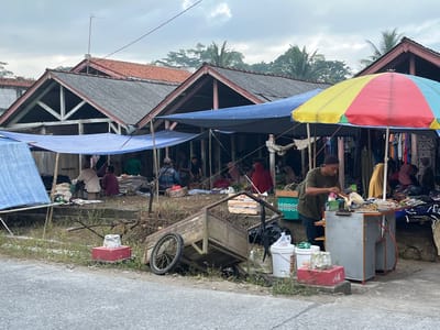 The image depicts an outdoor market scene in Tambakreja, Indonesia. Several stalls are set up under blue and colorful canopies. In the foreground, a man in a green shirt and dark cap is preparing food at a stand. Next to the stand are several buckets and a red platform. A wooden cart with large wheels is positioned on the side of a gray paved road. In the background, people are seated and interacting within the market stalls, which are lined with goods, including clothing. The architecture of the market stalls features pitched roofs with exposed wooden beams. The sky is overcast with some visible trees and palm fronds in the distance.