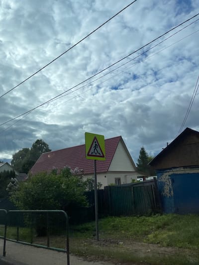 The photograph appears to be taken in Semyonovskoye, Russia. It shows a pedestrian crossing sign against a backdrop of a cloudy sky and houses. Power lines are visible in the foreground. There is vegetation and a fence on the right side of the image. The buildings have varied designs and colors. The overall impression is a quiet residential street scene.