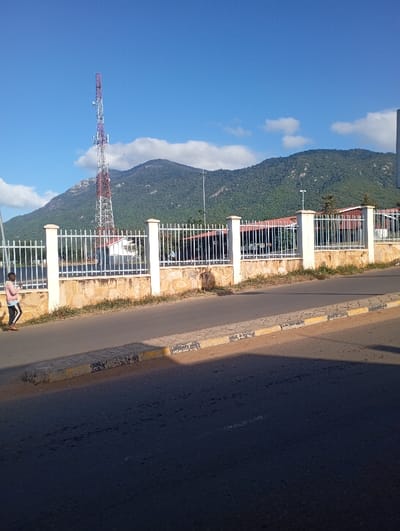 The image depicts a bright, sunny outdoor scene in Namanga, Kenya, featuring a road, a fenced property, and a mountainous backdrop. In the foreground, an asphalt road displays prominent shadows, suggesting either morning or late afternoon sunlight. A concrete curb, marked with yellow and black stripes, borders a sidewalk on the left side of the road.
A single person is visible on the far left, walking away from the viewer on the sidewalk. They are dressed in a light pink long-sleeved shirt, dark trousers, and light-colored shoes. Adjacent to the sidewalk stands a low stone wall, topped with a white metal picket fence supported by white pillars. Behind this fence, sections of a building with a distinctive red roof are visible.
The background is dominated by a majestic range of green, tree-covered mountains rising under a clear blue sky with scattered white clouds. A tall, red and white lattice communication tower stands prominently against the mountain on the left, serving as a notable landmark. Several streetlights are also discernible in the mid-distance, interspersed among the trees on the mountain slopes. The scene appears peaceful and devoid of any visible text.