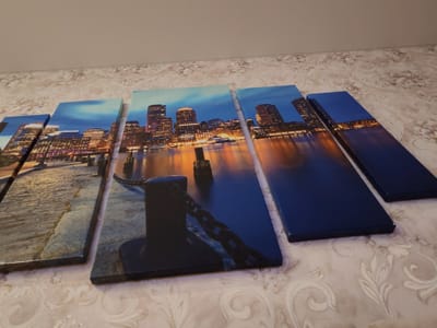 The image displays a multi-panel canvas art depicting a nighttime city skyline with illuminated buildings and their reflections on the water. The art is arranged in a sequence of vertical panels, with the leftmost panel showing a cobblestone path. The subsequent panels reveal a waterfront with a bollard and chains, followed by a wider view of the city's illuminated skyscrapers. The art is placed on a patterned surface, possibly a table or bed, and is leaning against a wall. The lighting in the scene is dim, suggesting an indoor setting with artificial light. There are no people or animals visible in the image. The artwork is the primary subject, with the background providing context for its display. The location of Kotovsk, Russia is not depicted in this image, as the artwork clearly shows a large, modern cityscape that does not resemble Kotovsk.