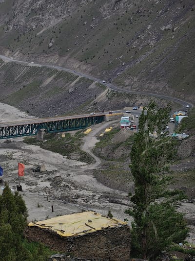 This image depicts a mountainous landscape in Darcha, India. The scene includes a winding road carved into the steep, rocky hillside, with several vehicles traveling on it. Below the road, a large, green metal truss bridge spans a dry riverbed. On the other side of the riverbed, near the bridge, a cluster of small buildings and tents are visible, possibly serving as a roadside settlement or rest stop. A tall, leafy tree stands prominently in the foreground, its branches swaying. In the lower left corner, a stone structure with a yellow tarp covering its roof is partially visible, suggesting a dwelling or storage area. Two flags, one red and one blue, are planted in the foreground. The overall impression is one of a remote, rugged, and challenging terrain. The weather appears overcast, with muted lighting conditions. The time of day is indeterminate due to the lack of strong shadows. The image conveys a sense of travel and human presence in a wild, natural environment.