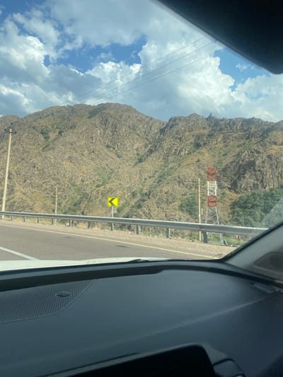 The image, taken from inside a vehicle, shows a scenic view of a mountainous landscape with a highway in the foreground. The mountains are rocky and rugged, with patches of green vegetation. A guardrail runs along the side of the highway. A yellow road sign with a black chevron pointing left is visible on a pole to the left of the road. Further along the road, a tall, red and white lattice tower stands next to a utility pole. The sky is partly cloudy with dramatic white clouds against a blue background. The interior of the car, including the dashboard and windshield, is visible at the bottom and top of the image, respectively. The location is identified as Saiyakka, Uzbekistan.