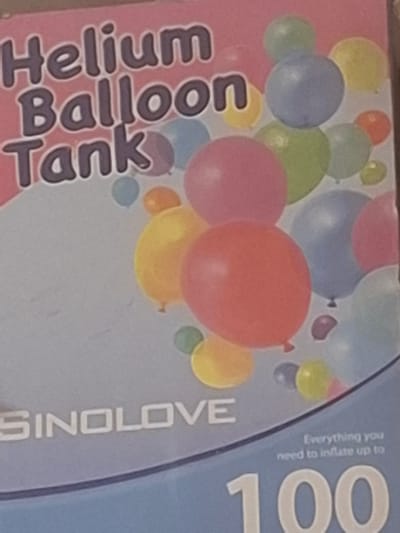 The image is a close-up, slightly blurry photograph of a pink and blue box that appears to be packaging for a helium balloon tank. The box has text that reads "Helium Balloon Tank" in large, dark purple, stylized letters. Below this, in white, is the brand name "SINOLOVE". Scattered across the box are various colorful balloons in shades of pink, blue, green, yellow, and orange. Towards the bottom right, there is smaller text that says "Everything you need to inflate up to" followed by a large "100". The background of the box is a gradient of light pink at the top to a light blue at the bottom. There is no visible indication of the city of Mıñbaev, Kazakhstan, in the image. The photograph is taken at a slight angle, suggesting it might be a product photo or a shelf display.
