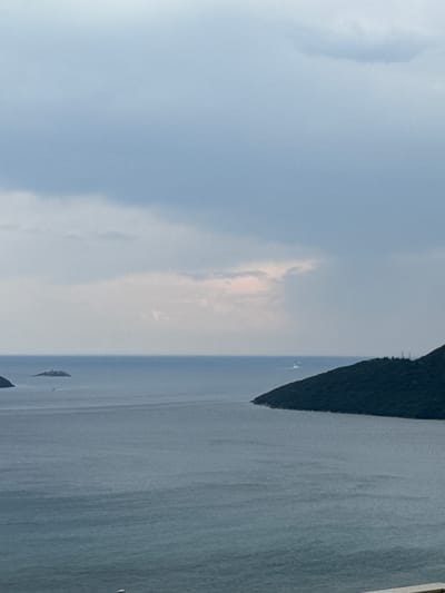 This is a landscape photograph of a body of water with a dark, forested landmass on the right side. The water is calm and appears to be a bay or a sea. There are small boats visible on the water. The sky on the left is filled with clouds, some of which are lighter and appear to be illuminated by sunlight, suggesting it might be daytime, perhaps late afternoon or early morning given the soft light. The landmass appears to be a steep hillside covered in dense vegetation. The overall impression is one of a serene and natural setting.