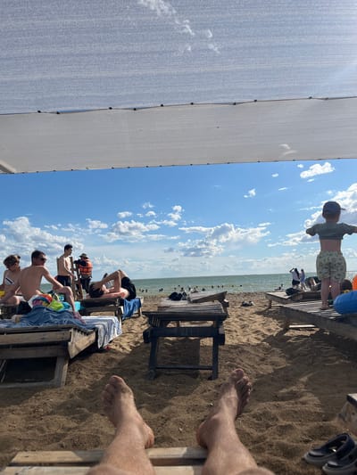 An eye-level, wide shot shows a sunny beach scene on a cloudy day. The foreground is dominated by a person's legs and feet, covered in sand, resting on a lounge chair. In the midground, several people are relaxing on lounge chairs, some are sitting up and chatting, while others are lying down. One person in an orange life vest stands by a ladder. Further in the background, people are visible in the water, swimming and wading. The beach is sandy, and the sea is calm. The sky is blue with scattered white clouds. The location is identified as the beach in Gres-2, Kazakhstan.