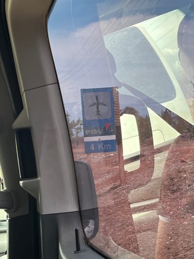The image depicts an exterior scene viewed from inside a vehicle, specifically through a window on the right side (likely the passenger side), in Las Bermúdez, Venezuela. The left portion of the frame is occupied by the vehicle's A-pillar and interior trim, composed of grey and dark grey plastic components. Reflections are prominent on the window surface, showing overhead power lines against a bright sky in the upper area, and the partial silhouette of a person's head and shoulder on the far right.

Outside the vehicle, a blue rectangular sign is visible on a brown post. The sign features a white silhouette of an airplane at the top, oriented towards the left. Below the airplane, the white capital letters "PDV" are displayed. Further down, a small red triangular arrow points towards the bottom left. The lowest section of the sign indicates "4 Km" in white text. The landscape behind the sign consists of reddish-brown, seemingly dry earth with sparse vegetation, including small, leafy trees or bushes. The ambient lighting suggests a bright, sunny day.