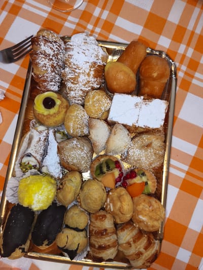 A rectangular golden platter of assorted pastries sits on an orange and white checkered tablecloth. The platter contains approximately two dozen individual pastries. At the top left are two large croissants dusted with powdered sugar. To their right are three small, golden-brown baked goods that appear to be rum baba. In the center of the platter, towards the top, is a round pastry with yellow cream and a dark cherry in the center. Below this are two cannoli, one filled with cream and pistachios, the other appearing to be empty or filled with a white substance. To the right of the cannoli are several small, puffy pastries dusted with powdered sugar, a rectangular pastry topped with powdered sugar, and a layered pastry. Towards the bottom left are two yellow, dome-shaped pastries. Below these are two rectangular pastries covered in dark chocolate glaze. Further to the right are several small, round, golden-brown pastries and two croissant-like pastries. In the bottom right section of the platter are two pastries that resemble braided bread. Several of the pastries have fruit toppings, including kiwi, berries, and apricot. A silver fork is visible on the tablecloth to the left of the platter. The background is blurred, but suggests an indoor setting with an orange and white checkered tablecloth. The scene is located in Campochiaro, Italy.