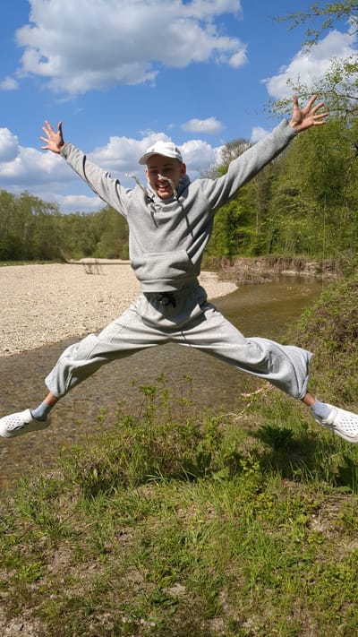 A young man is captured in a dynamic pose, appearing to jump or balance with his arms outstretched and legs spread, across a river and its bank. He wears a light grey hooded sweatshirt, matching grey sweatpants, a white baseball cap, and white clogs with light blue socks. He is smiling broadly, looking towards the camera, conveying a sense of joy and playfulness.
The setting is a vibrant outdoor natural environment, consistent with Beregovoe, Russia. The upper portion of the image shows a clear blue sky dotted with fluffy white clouds, indicating bright, sunny weather. Below, a shallow river with clear, light brown water flows. One side of the river features a wide bank covered in light-colored gravel, while the other side, further in the background, is lush with green trees, bushes, and grass. The time of day is bright, likely late morning or early afternoon, with natural light illuminating the scene. No visible text is present in the image.