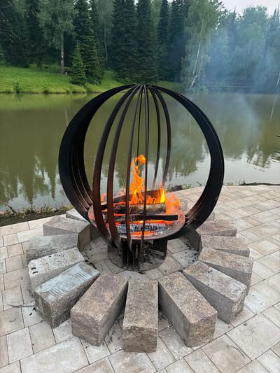 An outdoor fire pit with a flaming fire sits on a paved area next to a body of water. The fire pit is a black metal sphere with vertical slats and a circular base. It contains burning logs and embers, with flames reaching upwards. Around the fire pit, several large, rectangular stones are arranged in a semi-circle on the paving stones. The paved area is composed of light-colored, square and rectangular stones. In the background, a body of murky green water reflects the surrounding trees. A lush green, grassy bank slopes down to the water's edge, topped by a dense forest of tall trees, including evergreens and deciduous trees. Wisps of smoke are visible rising from the fire and drifting over the water. The sky appears overcast. The location is Staroabashevo, Russia.
