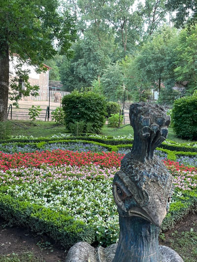 The image depicts an outdoor scene in the Nyíregyháza Animal Park, also known as Sóstó Zoo. In the foreground, a weathered, abstract sculpture of a bird's head, likely a peacock given the textured crown-like element, is prominently displayed. The sculpture is dark blue and grey, with visible texture suggesting it's made of stone or a similar material. Behind the sculpture, a meticulously maintained flower bed displays rows of vibrant flowers in shades of red, pink, white, and purple, bordered by neatly trimmed green hedges. In the middle ground, an elephant is visible in an enclosure, characterized by a fence and a light-colored building in the background. The background consists of lush green trees, indicating a natural or park-like setting. The overall atmosphere is one of a well-kept zoo or botanical garden.