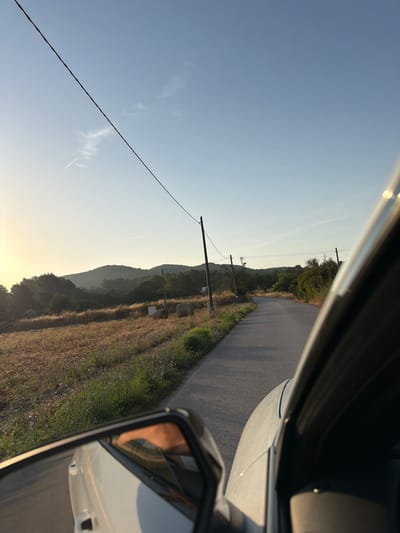This is a forward-facing, outdoor, long shot photograph taken from a vehicle, likely driving on a rural road in the municipality of Sant Joan de Labritja, Spain. The sun is low in the sky, casting a warm, golden light, suggesting either sunrise or sunset. The sky is a clear, pale blue with a faint contrail from an airplane visible in the upper left quadrant. A single power line stretches diagonally across the upper portion of the frame.
In the foreground, the right side of a white car is visible, including the A-pillar and a side mirror. The side mirror reflects a person's arm and hand. The road ahead is paved and curves gently to the right. On the left side of the road, a field of dry, golden grass extends towards rolling, tree-covered hills in the middle ground. Two cylindrical hay bales are visible in this field, along with several square, rough-hewn stones. Scattered among the grass are small, purplish wildflowers. Wooden utility poles with electrical wires are intermittently spaced along the right side of the road, receding into the distance. The overall environment appears serene and pastoral, characteristic of the rural landscapes found in Sant Joan de Labritja.