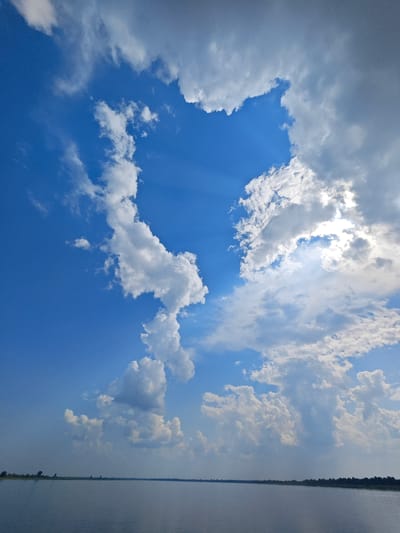The image depicts a vast, calm body of water, likely a river or lake, stretching towards a distant horizon. The water is a deep blue-grey, reflecting the sky above. Along the far bank, a thin line of dark green vegetation, possibly trees or reeds, marks the edge of the land. The sky is a brilliant blue, dominated by a dramatic display of white and grey clouds. Sunlight pierces through a gap in the clouds, casting visible rays downwards, illuminating the scene with a soft, ethereal glow. The clouds are varied in texture and form, some dense and voluminous, others wispy and translucent. The overall impression is one of natural beauty and tranquility. There are no people or man-made structures visible in the image, with the exception of a subtle, dark line on the horizon that might represent distant land features. The scene is consistent with a natural landscape in the city of Erkalovo, Russia, given the presence of large bodies of water and a temperate climate that supports such cloud formations.