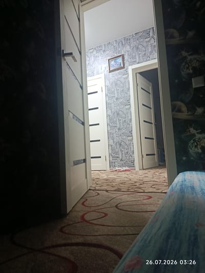 The image captures an interior hallway with two open doors. The left door is ajar, revealing a dark, heavily shadowed wall. The door on the right is fully open, leading into a room with patterned wallpaper and another open doorway further down. The hallway floor is covered in a carpet with a repeating circular design in red and brown. A framed picture hangs on the wall to the left of the right doorway. In the foreground, a person's leg, covered by a blue and white patterned fabric, is visible. A timestamp in the bottom right corner reads "26.07.2026 03:26". The location is identified as Oblivskaya, Russia.