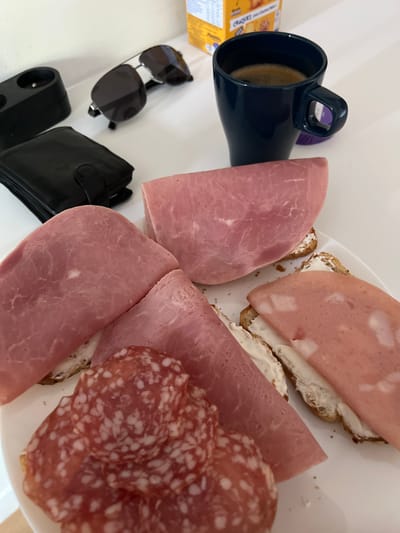 The image displays an overhead view of various items arranged on a clean white surface, likely a table. The central focus is a casual meal setup, primarily featuring a plate laden with deli meats and open-faced sandwiches.
On a white plate, several slices of pink deli meat, possibly ham or mortadella, are arranged, some folded and others topping small sandwiches. These sandwiches are made with brown bread, spread with a white creamy substance that resembles cream cheese. Additionally, there are multiple slices of red salami, distinctively dotted with white specks. Small crumbs are visible on the plate, suggesting it has been used.
To the left of the plate, a dark blue mug with a handle holds a light brown beverage, likely coffee or tea. Further to the left, a yellow and orange cardboard box is partially visible, with text fragments that appear to say "CHEES" and "SNACKS," indicating a product like crackers or cheese snacks.
Below these items, a pair of black sunglasses with dark lenses and silver accents is folded. Adjacent to the sunglasses is a closed black wallet. At the bottom left, a black electrical outlet extension or adapter with two sockets is present.
The scene suggests a daytime meal or snack, possibly breakfast, in an indoor setting such as a home or office. No people are visible in the image, and no specific location cues are discernible to confirm Vitry-sur-Seine, France.