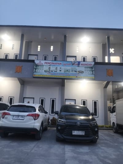 This media file depicts the exterior of a lodging facility at dusk. The building, identified by a sign as "PENGINAPAN Syariah UA MANDIRI," appears to be a hotel or guesthouse. The architecture is modern, with a multi-story facade featuring numerous windows and covered walkways. Two cars are parked in front of the building: a white Honda HR-V and a black Toyota Avanza. A white truck is partially visible on the right side of the frame.

The lighting suggests it is either early morning or late evening, with artificial lights illuminating the building. The sky is a dusky blue, indicating the transition between day and night. The ground appears to be a paved surface, possibly a parking lot or courtyard.

A prominent sign on the building displays the name of the establishment in large letters and includes smaller text with an address: "J. Bireuen - Takengon Ds. Lut Kucak Kec. Wih Pesam Kab. Bener Meriah Prov. Aceh." Additionally, the license plates of the vehicles are visible: the white car has plate "B 1338 FRU" and the black car has plate "BK 1595 AD0."

There are no people prominently featured in the image, and the scene conveys a sense of quietness and readiness for guests. The overall impression is that of a functional accommodation establishment in Indonesia, situated in a locale with a clear address.