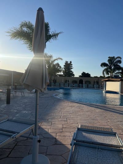 A sunlit outdoor swimming pool area is captured in the late afternoon, bathed in the golden light of a setting sun. The pool, with its clear blue water, curves gently through the scene. Several lounge chairs are visible, some folded and stacked, others set up on the stone-tiled patio. A closed umbrella stands prominently in the foreground, casting a long shadow. Palm trees and other vegetation surround the pool area, contributing to a tropical ambiance. In the background, a building with arched openings and a small bar structure are visible. The sky is a clear, brilliant blue. The scene appears to be a resort or hotel property. The location is the city of Otranto, Italy.