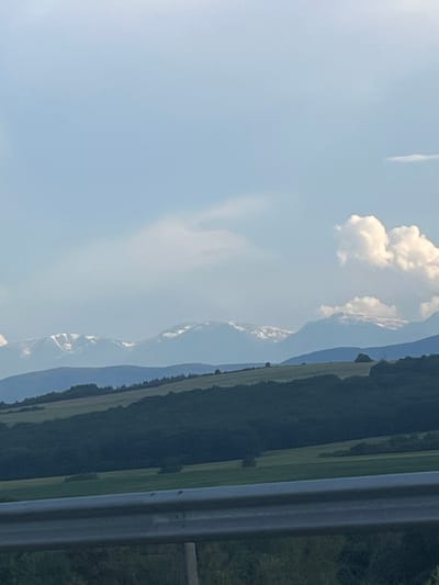 The image shows a natural landscape with rolling hills and mountains in the distance. In the foreground, there is a metal railing, suggesting a viewpoint or a roadside. The hills are covered with dark green trees and lighter green fields, indicating a mix of forest and agricultural land. The mountains in the background are capped with snow, contrasting with the verdant lower slopes. The sky is partly cloudy with a light blue hue. The image appears to be taken on a bright, possibly overcast day. No people or specific landmarks are visible, and it is not possible to determine the exact location in Vakarel, Bulgaria, from the image alone.