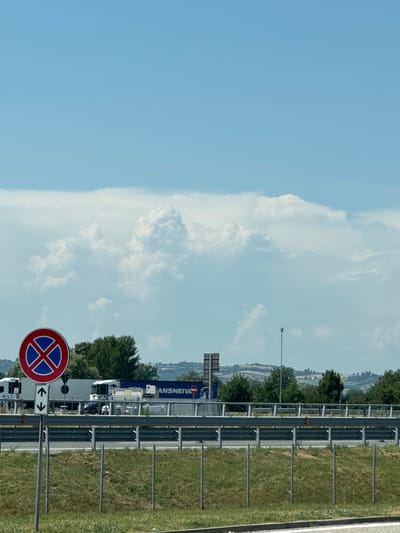 The image shows a roadside scene on a sunny day, likely in or near Occimiano, Italy, given the context of its metadata. In the foreground, a grassy embankment is bordered by a metal guardrail. To the left, a traffic sign with a red circle and a blue "X" indicating no stopping is prominently displayed, accompanied by a smaller sign with vertical arrows pointing up and down.
Behind the guardrail, a road is visible with several vehicles. A large white truck with a blue stripe and the partial word "ANSNEIVA" on its side is parked or stopped. A smaller white vehicle is positioned behind it. Further in the background, a line of trees forms a natural barrier, with rolling hills and some buildings visible in the distance. The sky is a clear blue, with large, puffy white clouds dominating the upper portion of the frame. A single street lamp stands to the right of the frame in the mid-ground. The overall impression is one of a typical highway or main road setting in a rural or semi-rural Italian landscape.