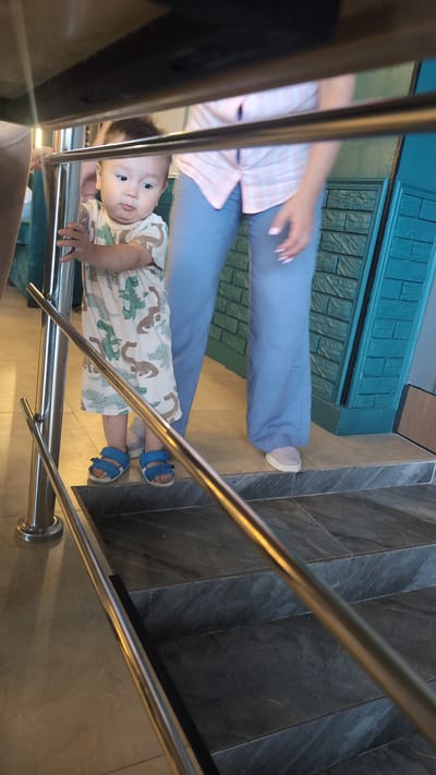 A toddler stands on a tile step, holding onto a metal railing. The toddler wears a light-colored shirt with a dinosaur pattern and blue sandals. Behind the toddler, a person in light blue pants and a pink and white plaid shirt is visible. The background shows a teal brick wall and a staircase with dark gray marble steps. The scene appears to be indoors, possibly in a restaurant or public building in Ötegen batyr, Kazakhstan.